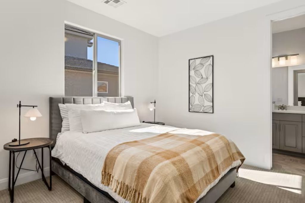 A bedroom with a bed, bedside tables, a window, a painting, and a bathroom visible in the background.