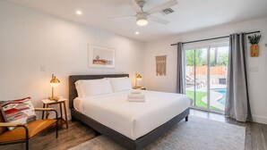 Quarto com cama, luminárias de cabeceira, uma cadeira, escrivaninha, ventilador de teto e vista para área de piscina.