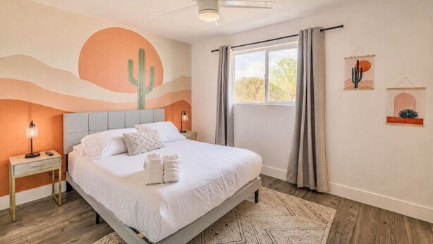 Quarto com cama, mesa de cabeceira, ventoinha no teto, janela com cortinas e decoração nas paredes.