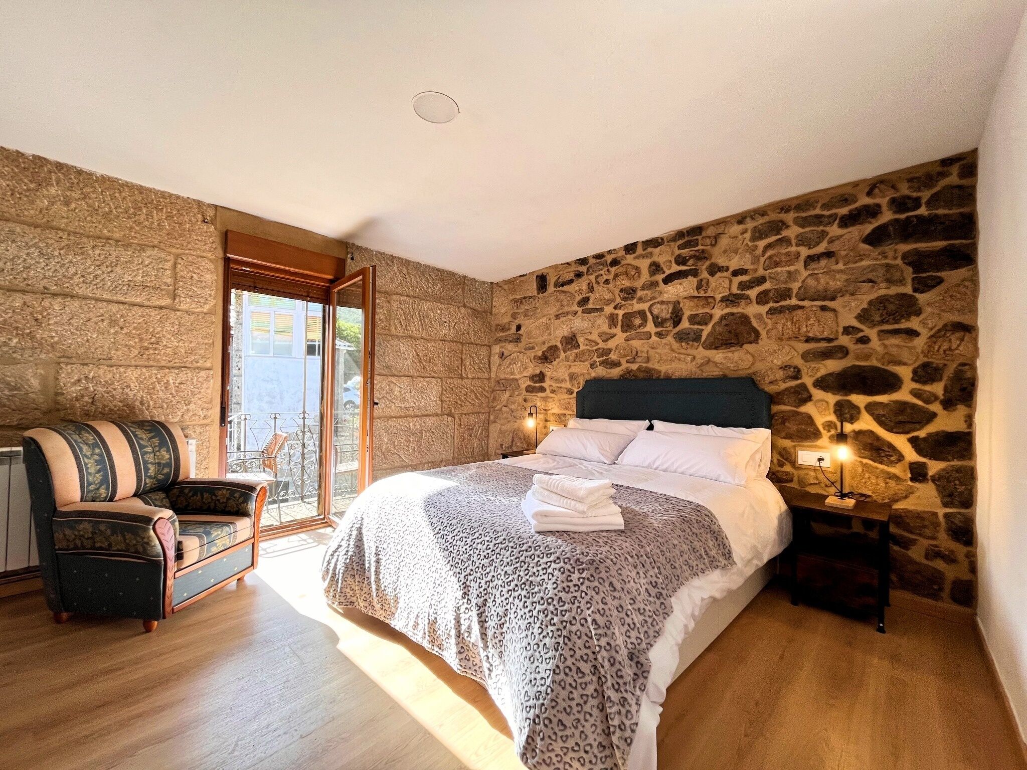 A bedroom with a stone wall, a bed with a leopard print blanket, a chair, and a nightstand with a lamp.