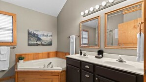 A bathroom with a wooden bathtub, two sinks, dark cabinets, a large mirror with lights, and a painting on the wall.