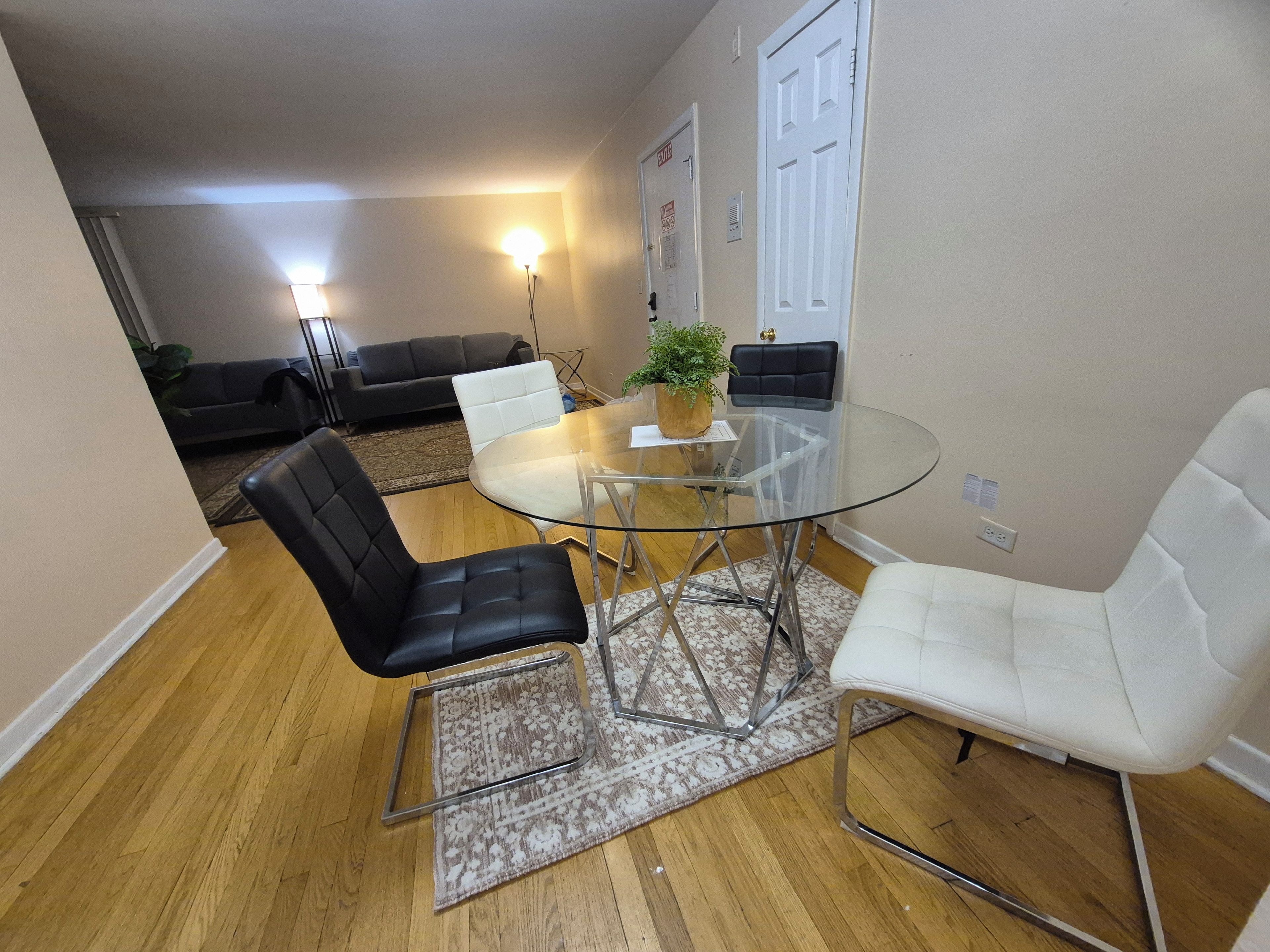 A dining area with a glass table, four chairs, a potted plant, and a sofa in the background.