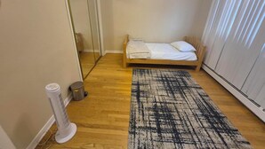 Ein kleines Zimmer mit einem Einzelbett, Holzboden, einem großen Spiegel, einem Heizlüfter und einem Fenster mit Jalousien.