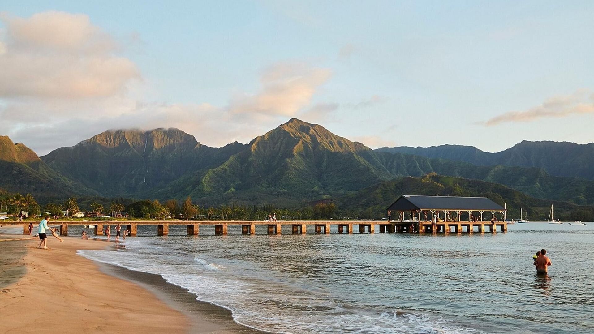 A beach with a pier extending into the water, a mountain range in the background, and a building on the pier.