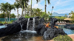 A pool area with a waterfall feature, surrounded by palm trees and a mountain view. - Princeville North Shore Escape - The Cliffs Club 1BR Suite, Ocean Area (Princeville)