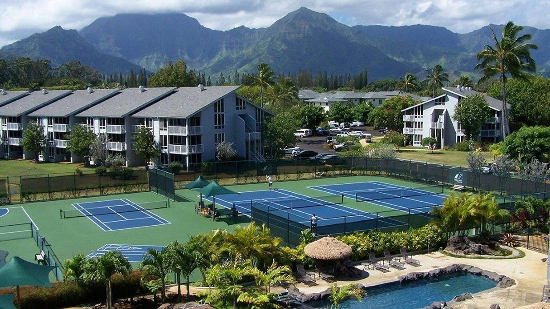 Un complexe de courts de tennis comprenant plusieurs terrains, entouré de bâtiments et avec une chaîne de montagnes en arrière-plan.
