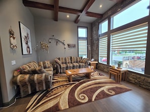 A living room with a sectional sofa, a coffee table, and a rug. There are windows with blinds, a stone wall, and artwork on the wall.
