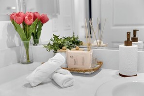Une salle de bain avec un lavabo blanc, un vase en verre contenant des fleurs roses, un plateau en bois avec des articles de toilette et une serviette blanche.