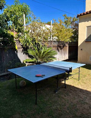 An outdoor ping pong table set up in a backyard with a wooden fence, trees, and a house in the background. - Spanish Gem in West Hollywood (Los Angeles)