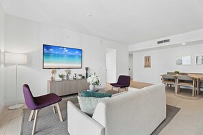 Ein modernes Wohnzimmer mit einem großen Flachbildfernseher, einer Couch, zwei Sesseln, einem Couchtisch und einem Essbereich mit Tisch und Stühlen.