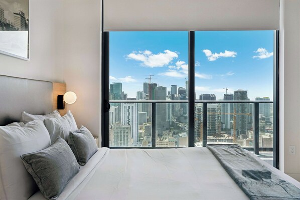 A bedroom with a large bed, a nightstand, a wall-mounted lamp, and a city view through floor-to-ceiling windows. - Paloma Place, Downtown Miami, Pool and Gym (Miami)