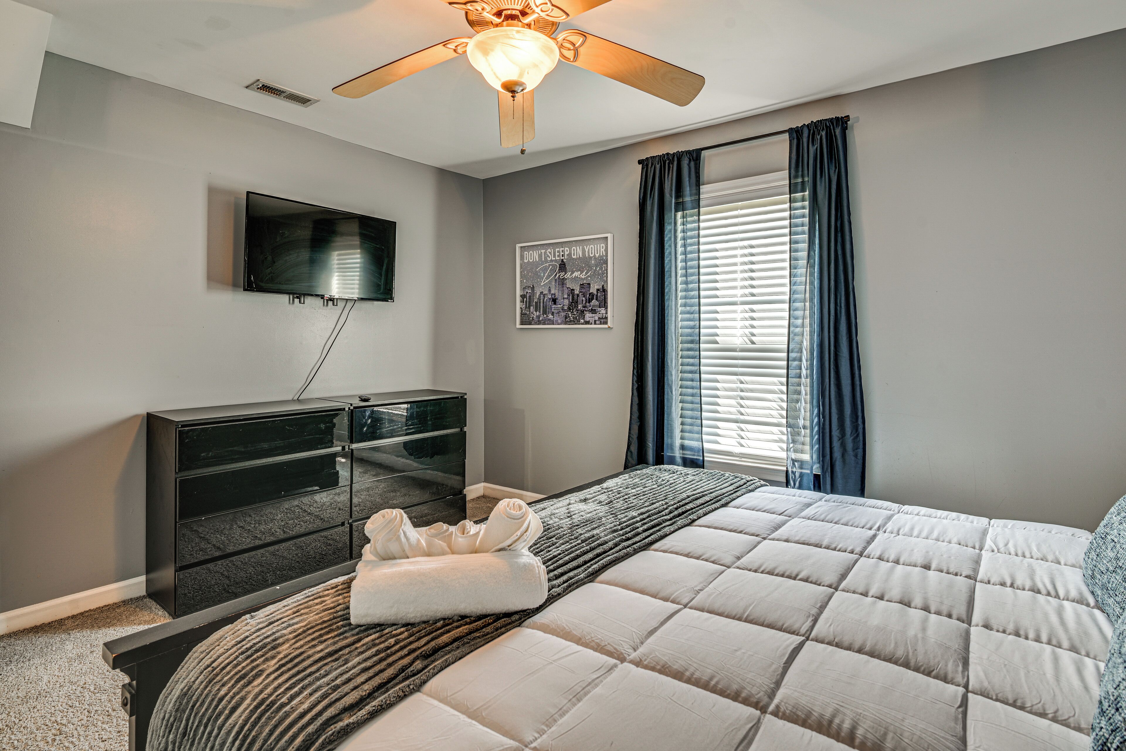 Une chambre à coucher avec un lit, un téléviseur fixé au mur, une commode et une fenêtre avec des rideaux.