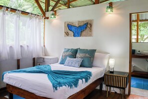 A bedroom with a bed, pillows, a nightstand, a wall art piece, and a window with curtains. - Oceanfront Open-Air Bungalow on Ballena Beach (Uvita)