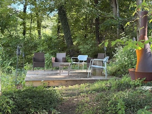 An outdoor deck with wicker furniture and a metal chair, surrounded by dense greenery.