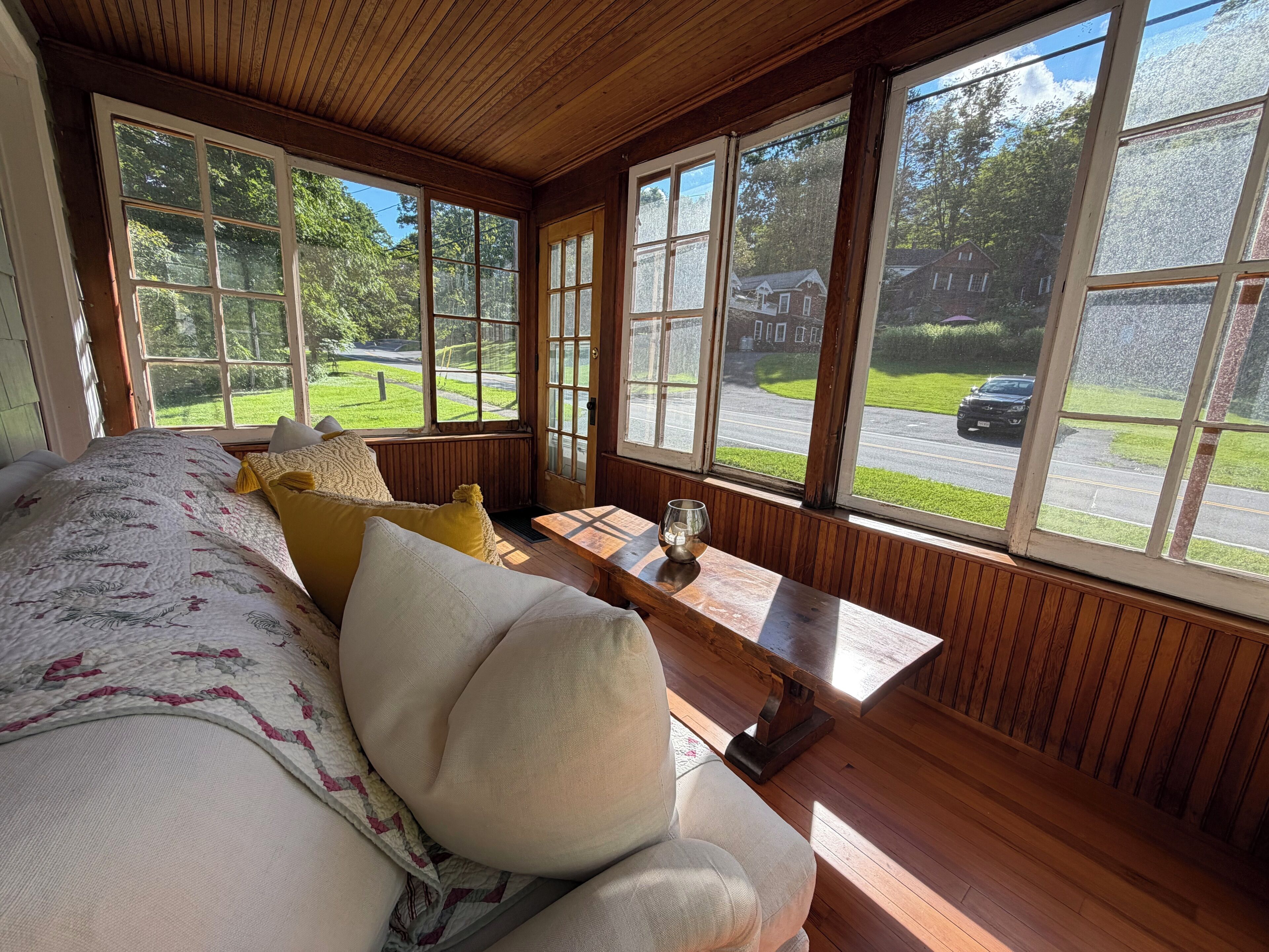 A room with a large bed, a wooden bench, and a view of a street and greenery.