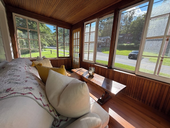 A room with a large bed, a wooden bench, and a view of a street and greenery.
