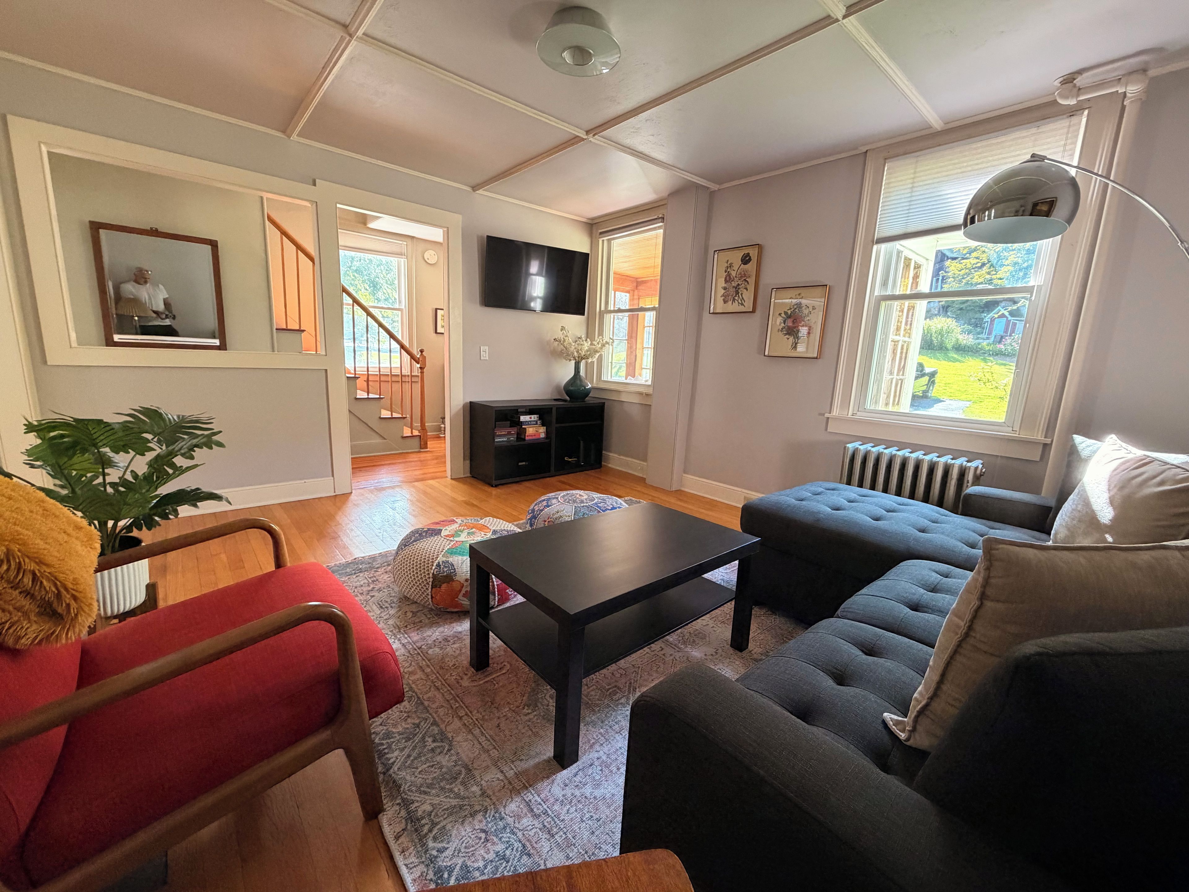 A living room with a sofa, armchair, and a coffee table. There is a staircase leading to another room, a TV mounted on the wall, and a window with a view of the outdoors.