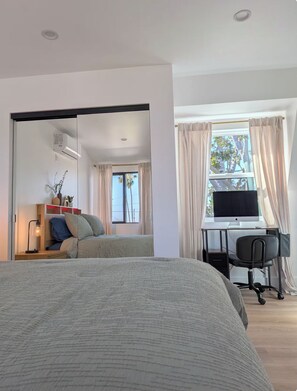 Ein Schlafzimmer mit einem Bett, einem Schreibtisch mit Computer, einem Stuhl und einem Fenster mit Vorhängen.