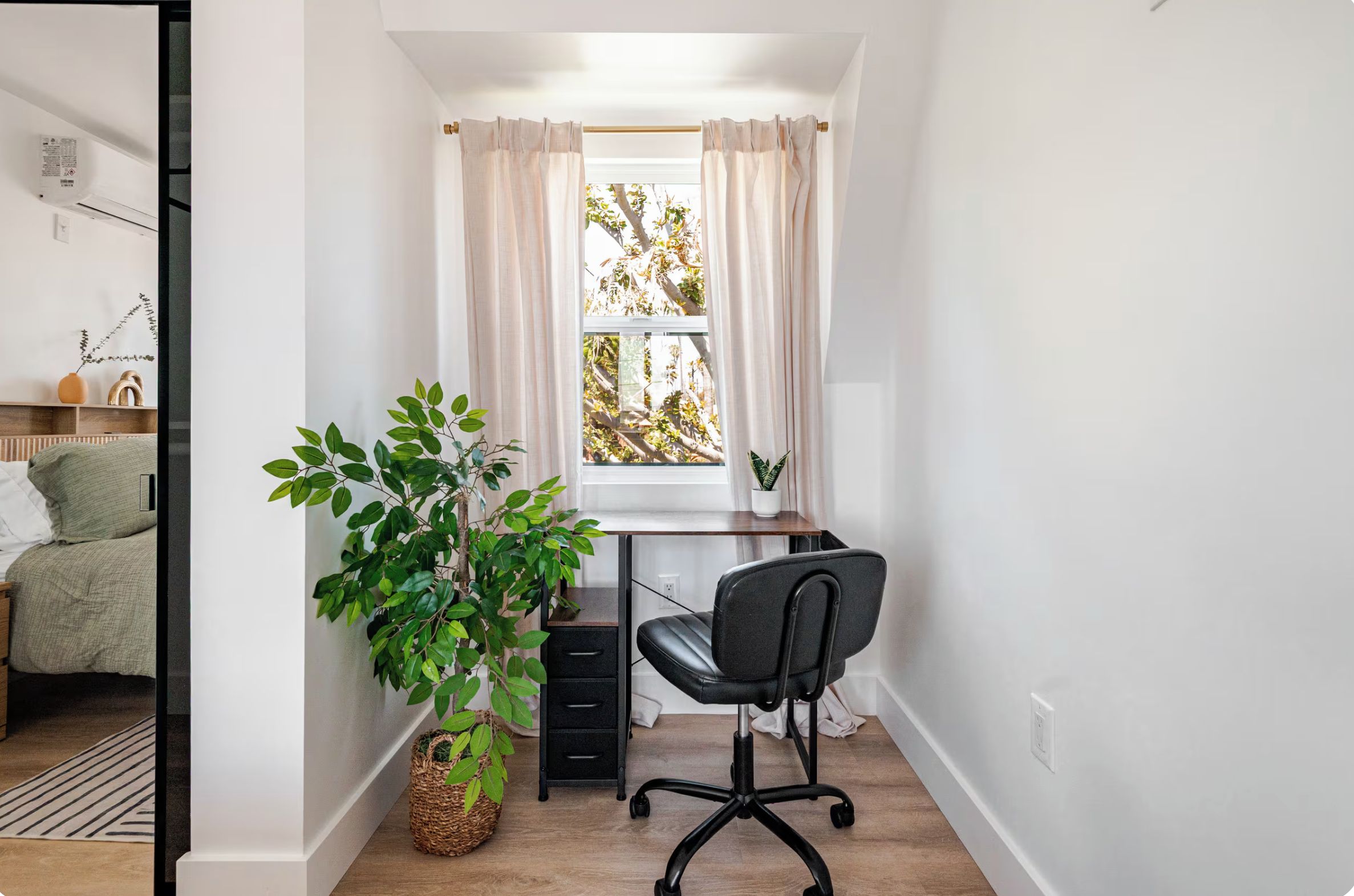 A small, well-lit room with a desk, chair, and a large window with curtains. There is a potted plant on the desk and a small plant on the windowsill.
