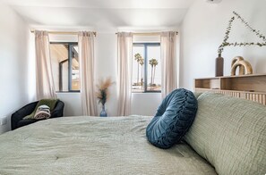 A bedroom with a large bed, a chair, a vase with dried plants, and a view of palm trees through the window. - Stylish 3BR Windsor Hills Retreat Near SoFi & LAX (View Park-Windsor Hills)
