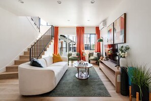 A modern living room with a curved sofa, a coffee table, and a staircase with orange curtains. - Stylish 3BR Windsor Hills Retreat Near SoFi & LAX (View Park-Windsor Hills)