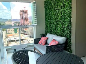 Ein Balkon mit Blick auf eine Stadtlandschaft, eine grüne Wand, ein graues Sofa mit roten Kissen und ein runder Tisch.