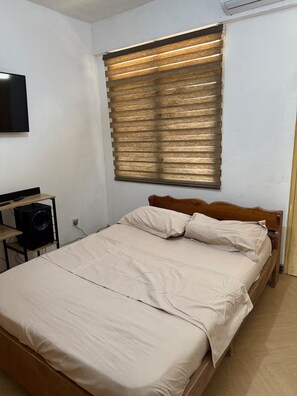 Ein Schlafzimmer mit einem Bett, einem an der Wand montierten Fernseher und einem Fenster mit Jalousien.