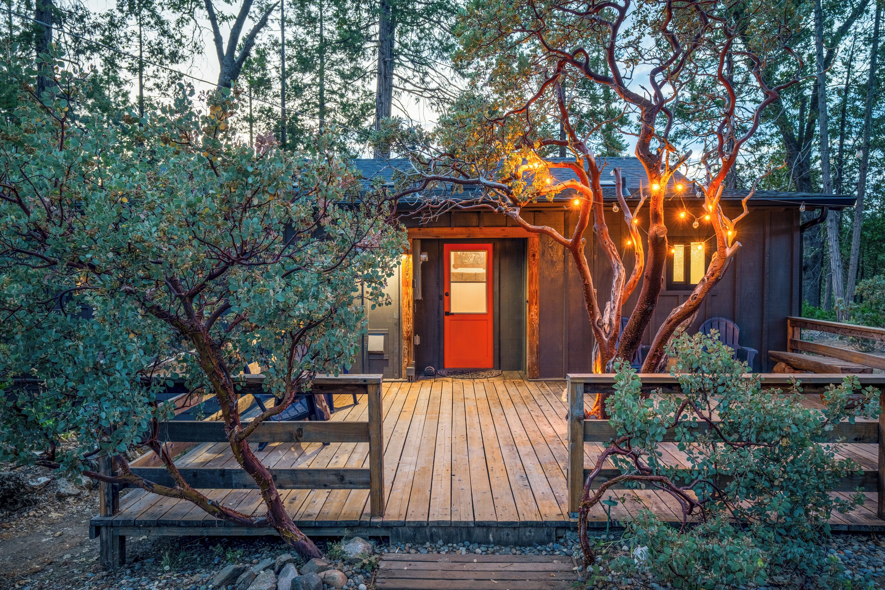 Una terraza de madera que conduce a una cabaña con puerta roja, rodeada de árboles y una valla de madera.