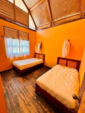 Een kamer met twee eenpersoonsbedden, een houten vloer, rieten dak en een raam met gordijnen.