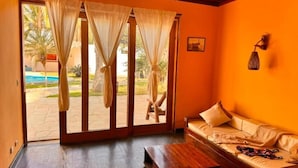 Ein Zimmer mit Holzboden, einer Couch mit Kissen, einem kleinen Tisch und Blick durch eine Glastür auf einen Außenbereich mit Pool und Palmen.