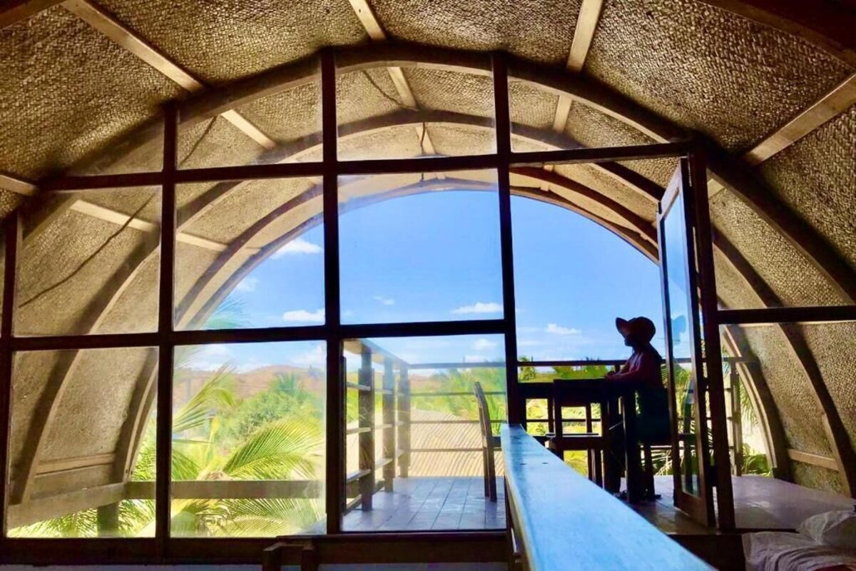 Een kamer met een gebogen plafond en grote ramen, met uitzicht op palmbomen en een heldere lucht.