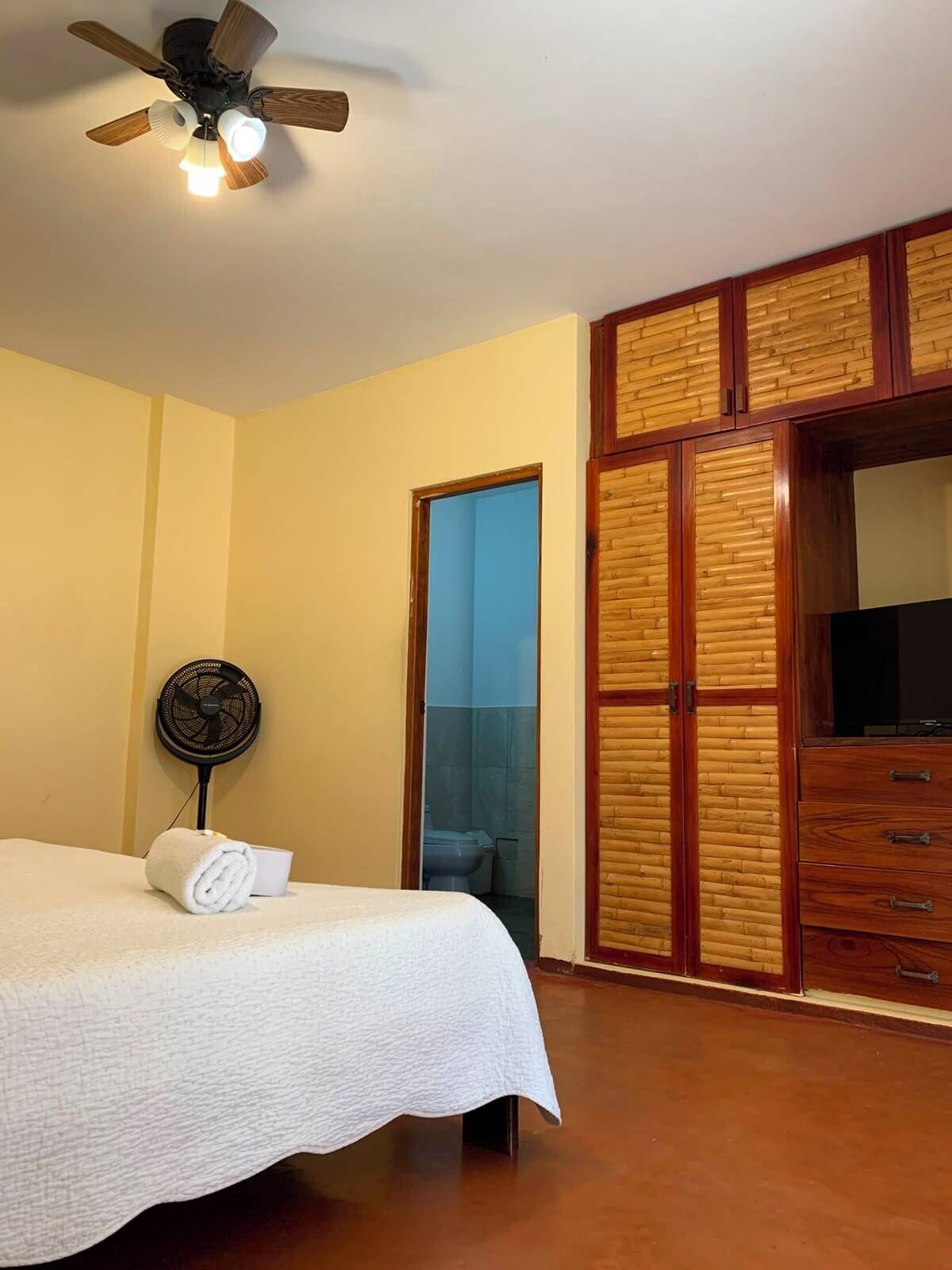 A hotel room with a bed, a ceiling fan, a wall-mounted fan, a wooden wardrobe, and a bathroom visible through an open door.
