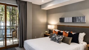 Habitación de hotel con una cama grande, una mesita de noche con una lámpara, vista a árboles a través de una ventana y un balcón con una silla.