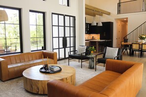 A spacious living room with leather sofas, a round coffee table, and a dining area with a kitchen in the background. - Maison Blanche I French-Inspired Retreat on 2.5 Acres I Sleeps 13 (Austin)