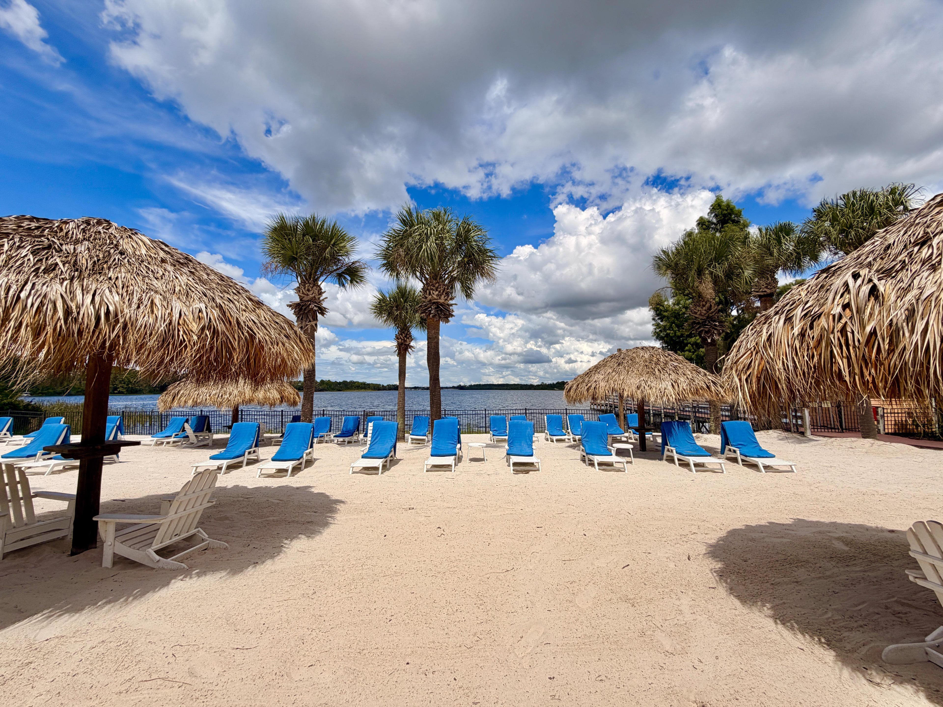 A beach with sun loungers and thatched umbrellas, palm trees, and a clear sky.
