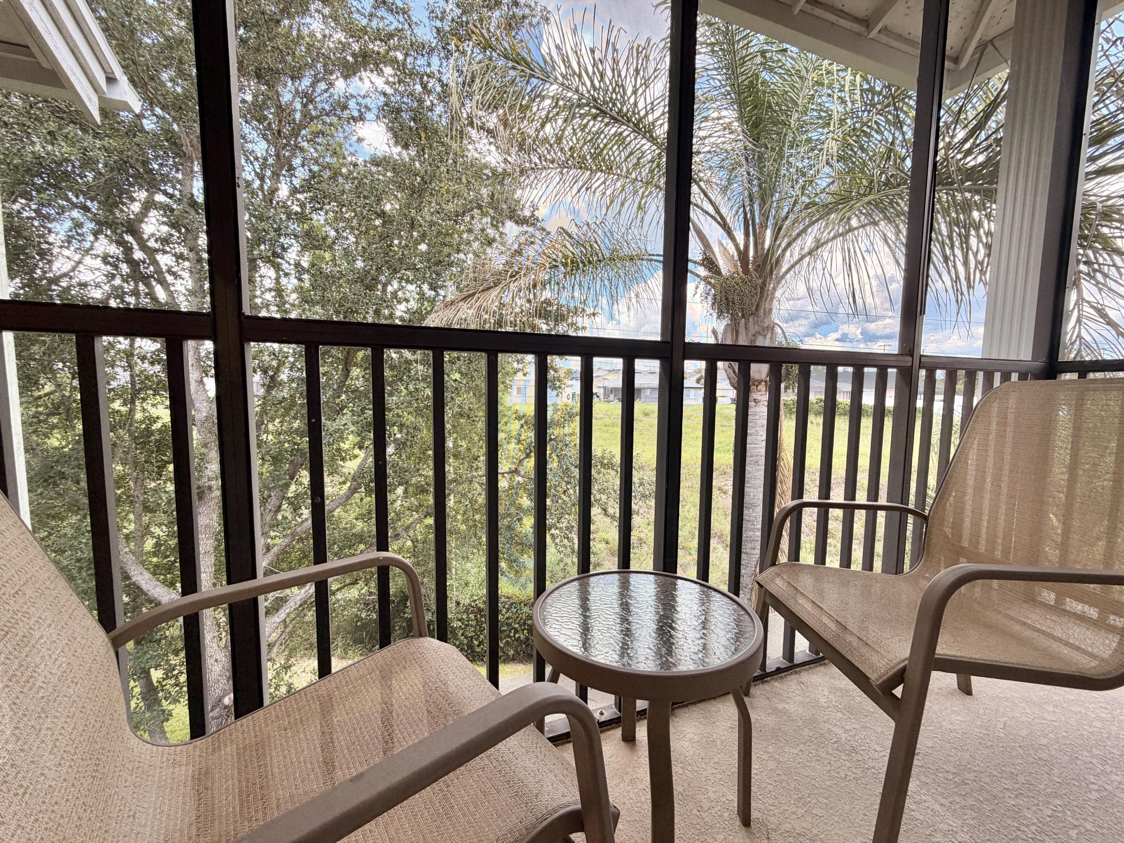 A balcony with a glass table and two chairs, overlooking a garden with trees and a palm tree.