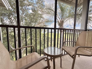 A balcony with a glass table and two chairs, overlooking a garden with trees and a palm tree. - 3BR Orlando Area Condo | Resort Pools & Pickleball (Davenport)