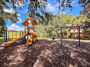 Un'area giochi con uno scivolo e altalene, circondata da alberi e un edificio sullo sfondo.