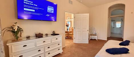 A hotel room with a large flat-screen TV displaying a welcome message, a white dresser with drawers, a bed with pillows, and a door leading to another room.