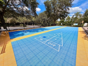 A shuffleboard court with a blue and yellow surface, surrounded by a fence and trees. - 3BR Orlando Area Condo | Resort Pools & Pickleball (Davenport)