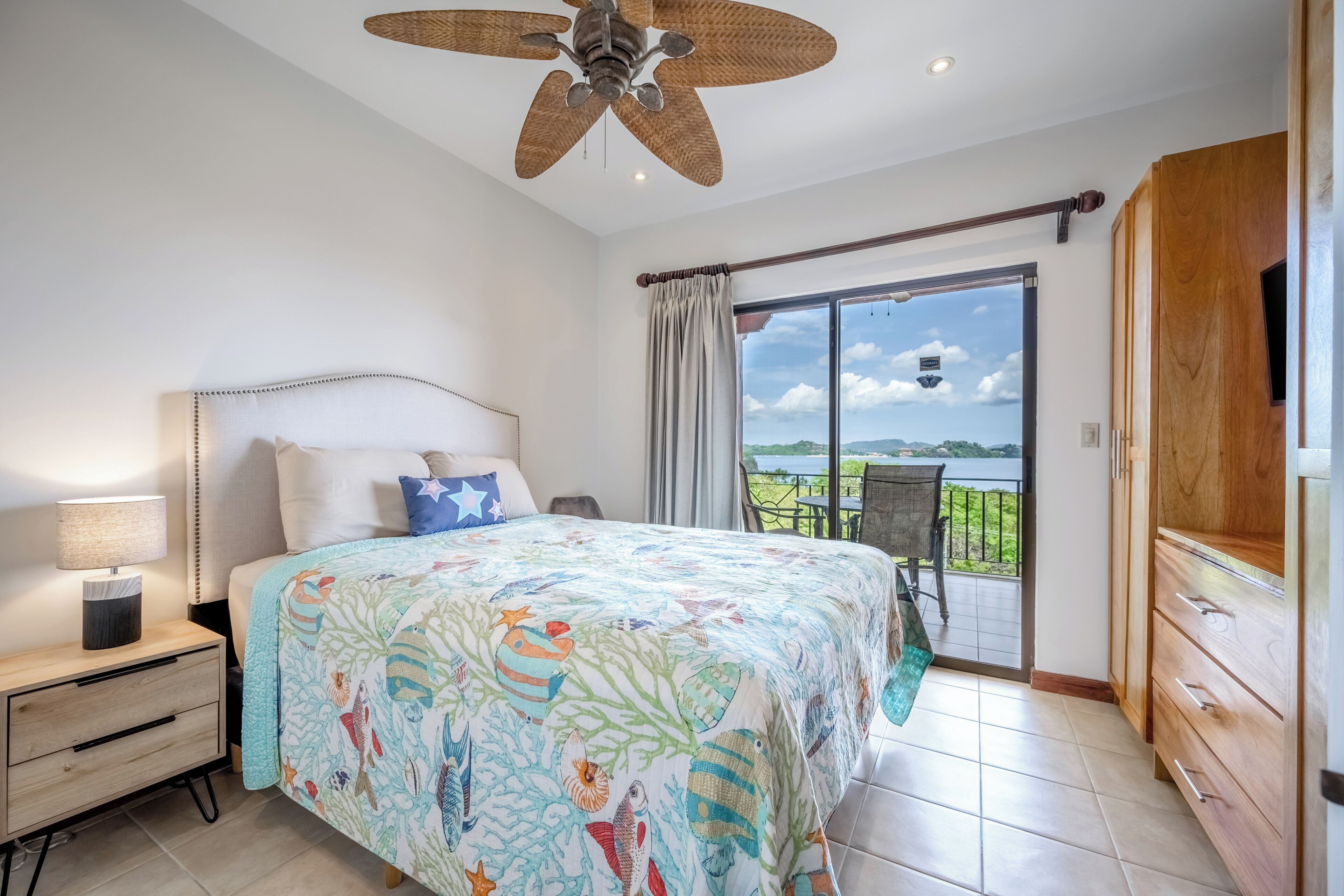 Quarto com cama, mesa de cabeceira, luminária e vista para uma varanda com mesa e cadeiras.