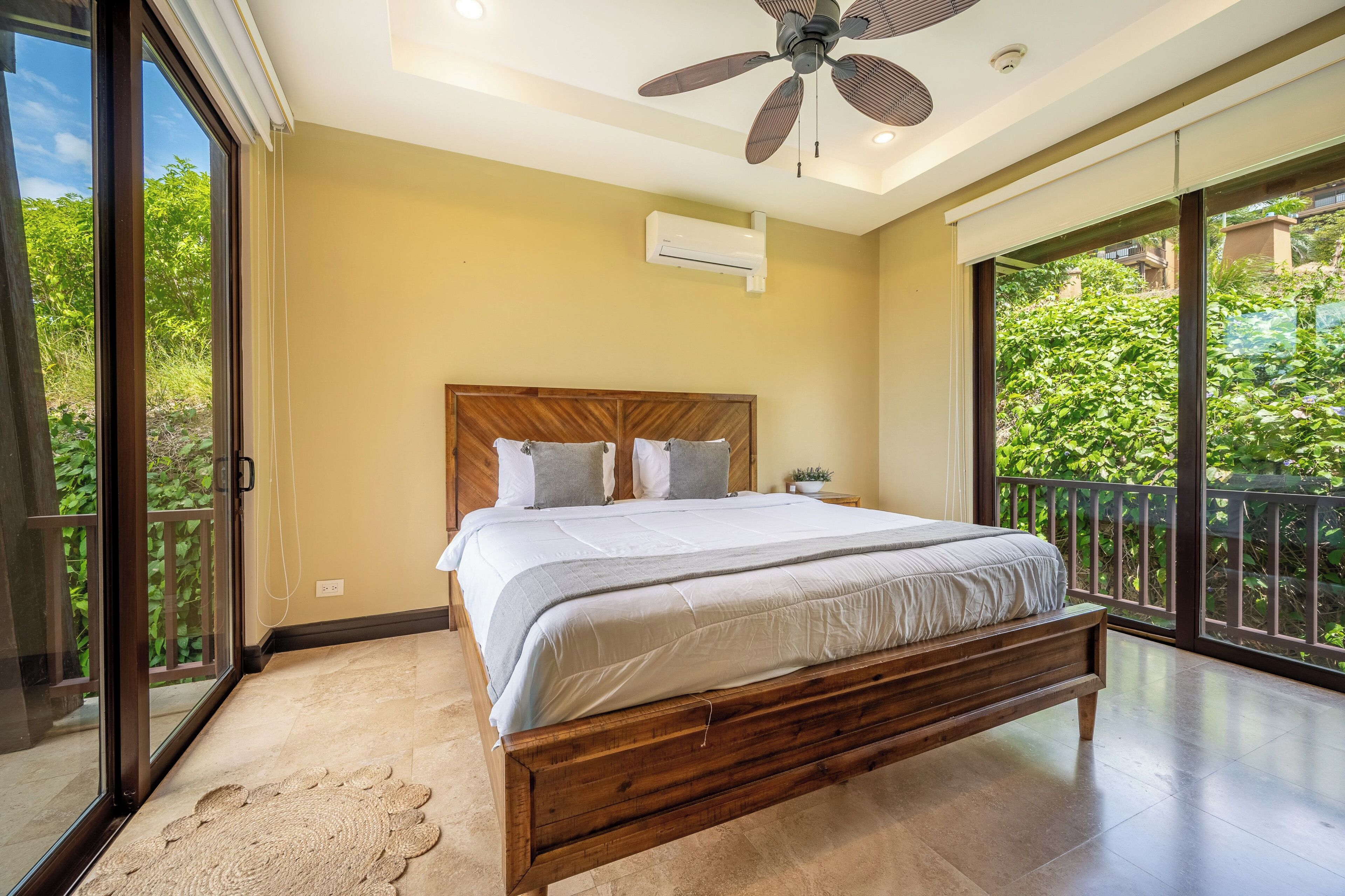 A bedroom with a large bed, a ceiling fan, and a view of greenery through the sliding glass door.