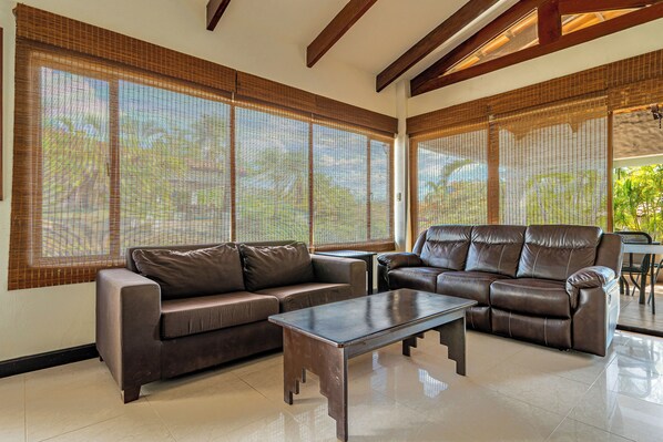 A spacious living room with a brown leather sofa, a matching armchair, and a coffee table. Large windows with bamboo blinds allow natural light to fill the room.