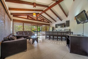 Amplia sala de estar con sofá, mesa de centro y mesa de comedor con sillas. Hay una zona de cocina con refrigerador, microondas y un televisor de pantalla plana montado en la pared.