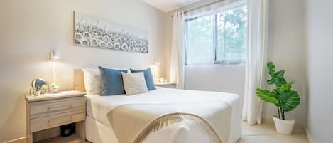 A bedroom with a bed, bedside tables, a wall-mounted artwork, a window with curtains, and a potted plant.