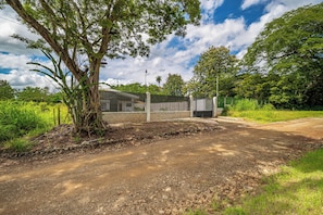 A rural road with a dirt path leading to a gated area with a brick wall and metal gate. - Stylish 'Casa Nonos' Home w/ Yard & Fire Pit! (Playas del Coco)