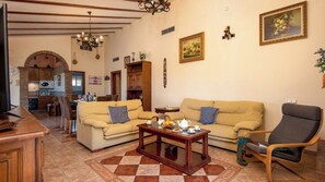 Ein gemütliches Wohnzimmer mit einer Couch, Sesseln und einem Couchtisch. Im Hintergrund ist eine Küche mit Essbereich zu sehen.