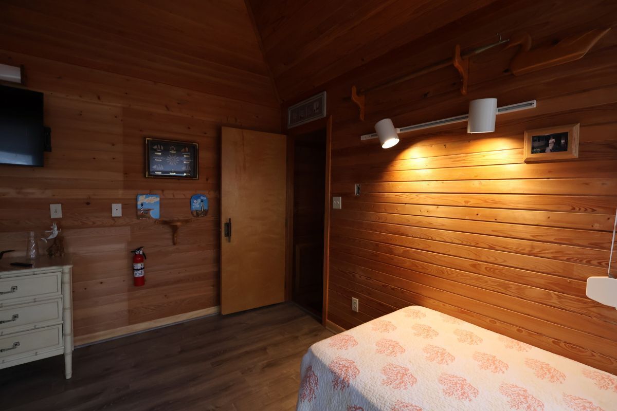 A wooden cabin room with a bed, a dresser, a mounted television, a fire extinguisher, and a wall-mounted thermostat.