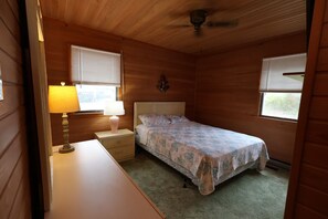 Une chambre avec un lit simple, une table de chevet, une lampe et un ventilateur de plafond.