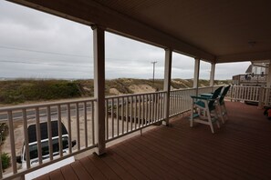 Een overdekt terras met een houten dek, witte leuningen en uitzicht op het strand en de duinen.